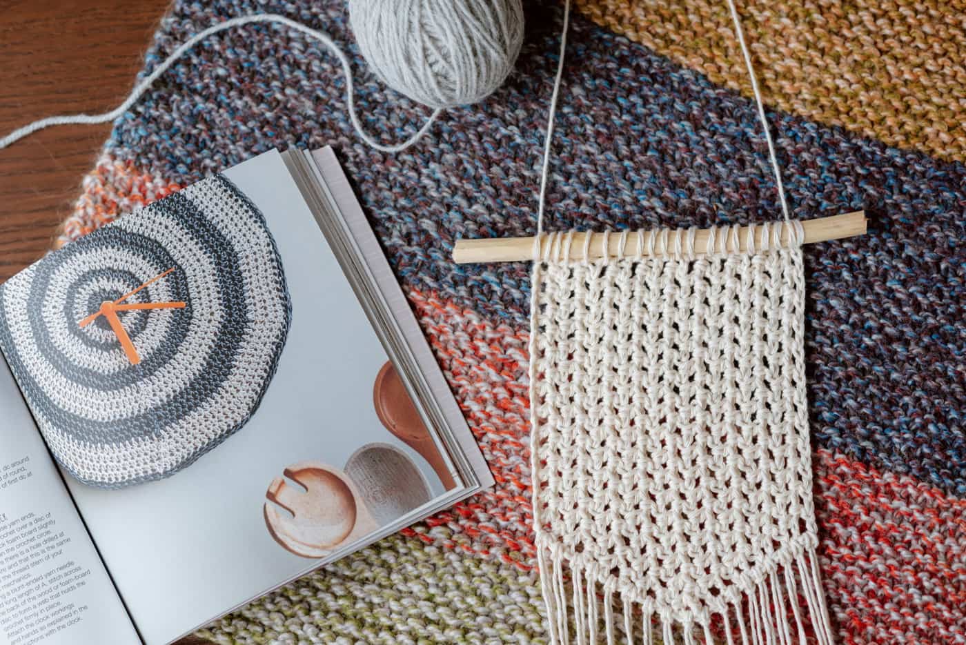 A striped knitted blanket with a ball of yarn, an open pattern book and a macrame hanging