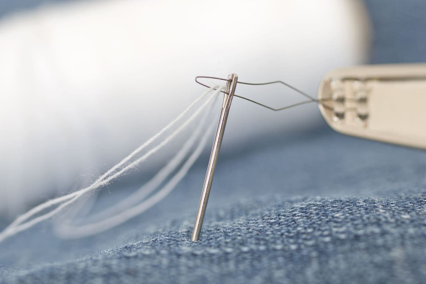 A macro photograph of a sewing needle threading white thread through denim fabric