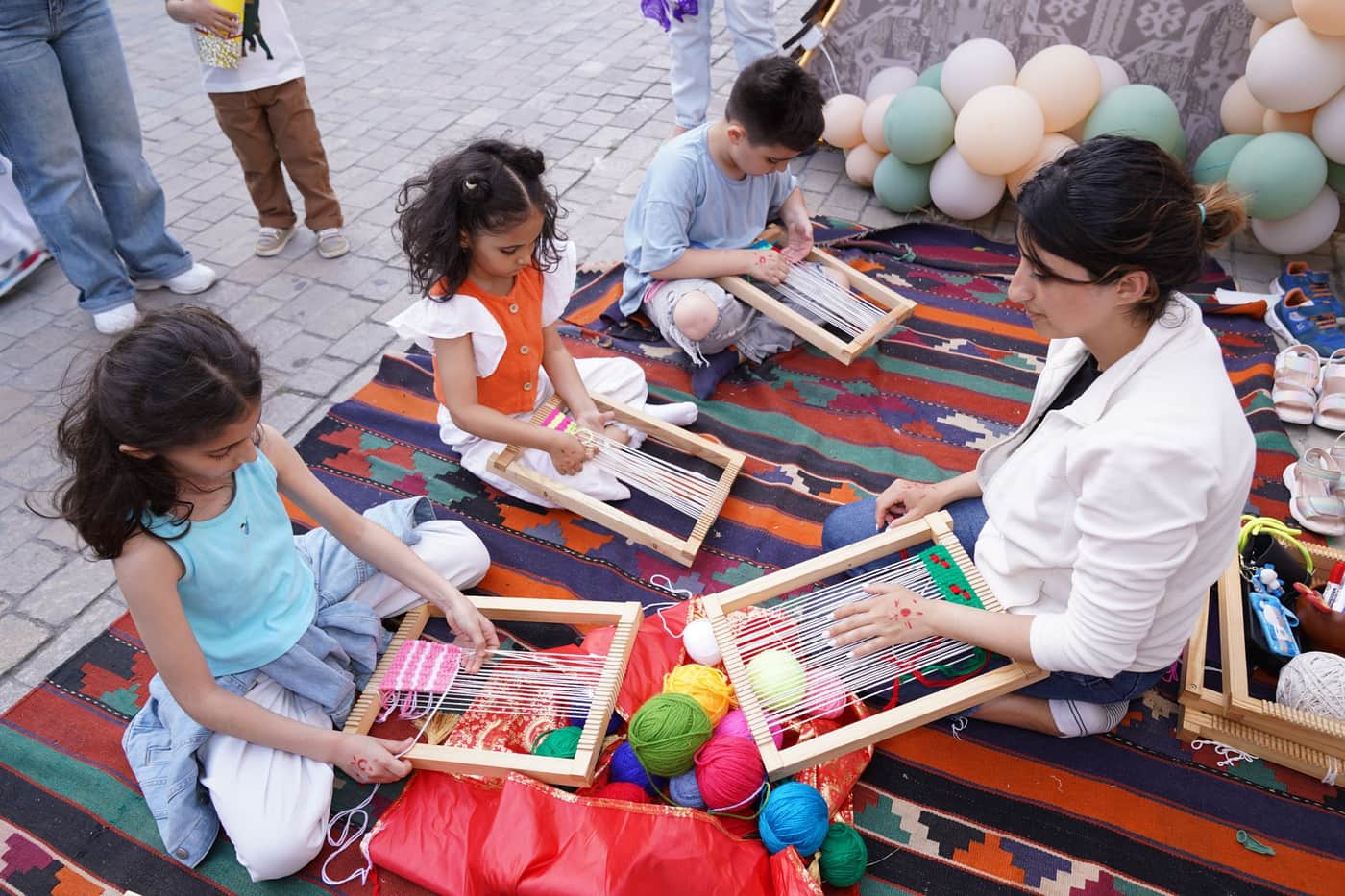 Children and an adult weaving together outdoors on a colorful mat surrounded by yarn