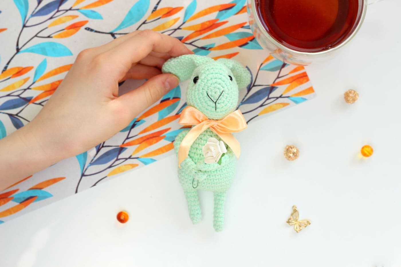 A hand holding a crocheted bunny toy beside a cup of tea on a decorated table