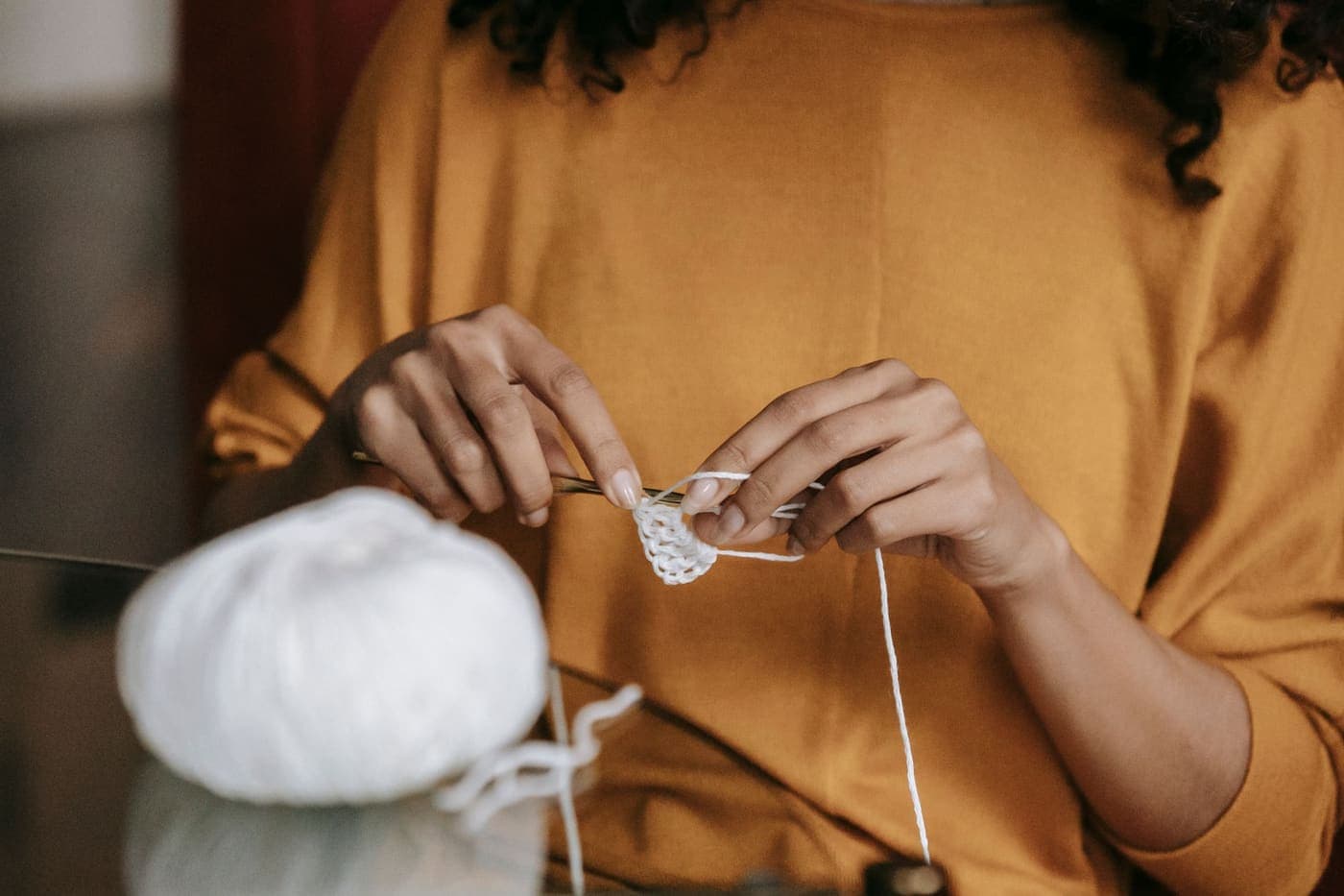 Hands crocheting with a hook and white yarn, a ball of yarn resting on the table