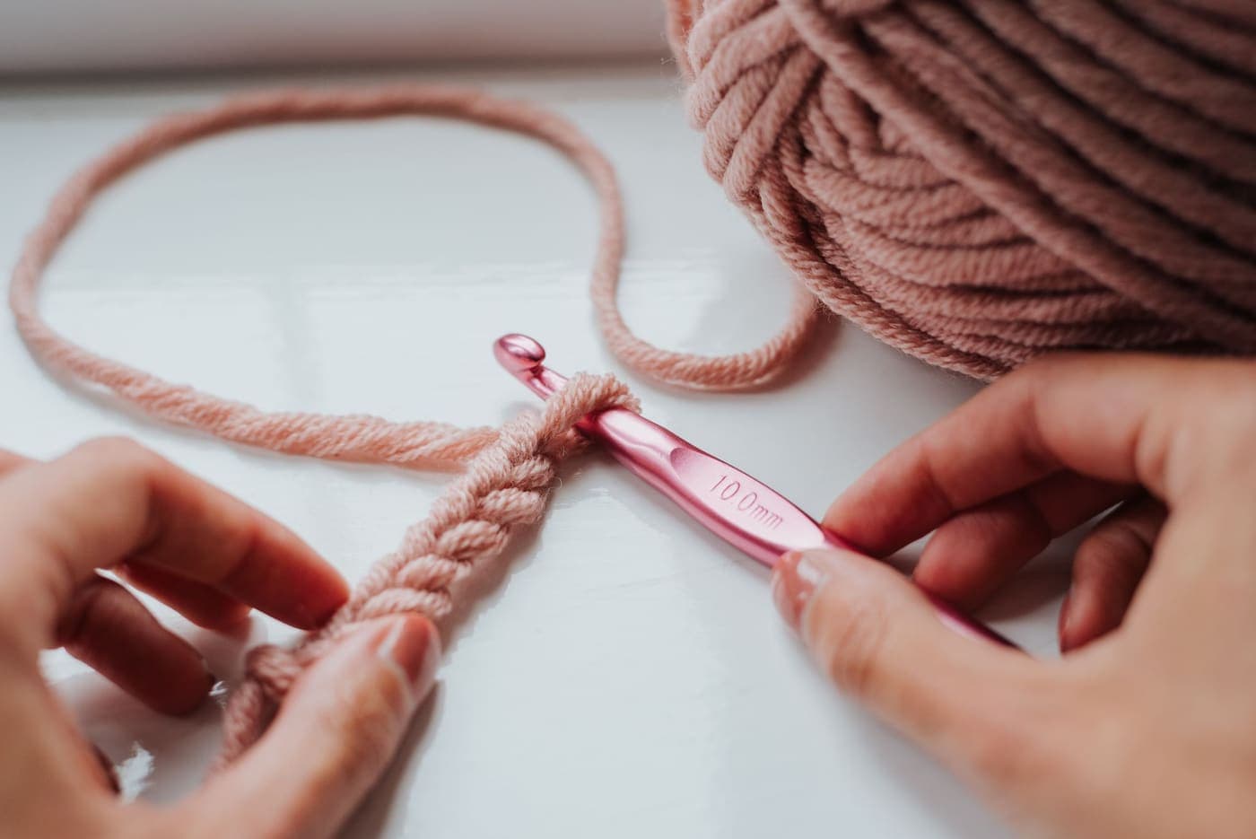 A person crocheting soft pink fabric with a hook by a window