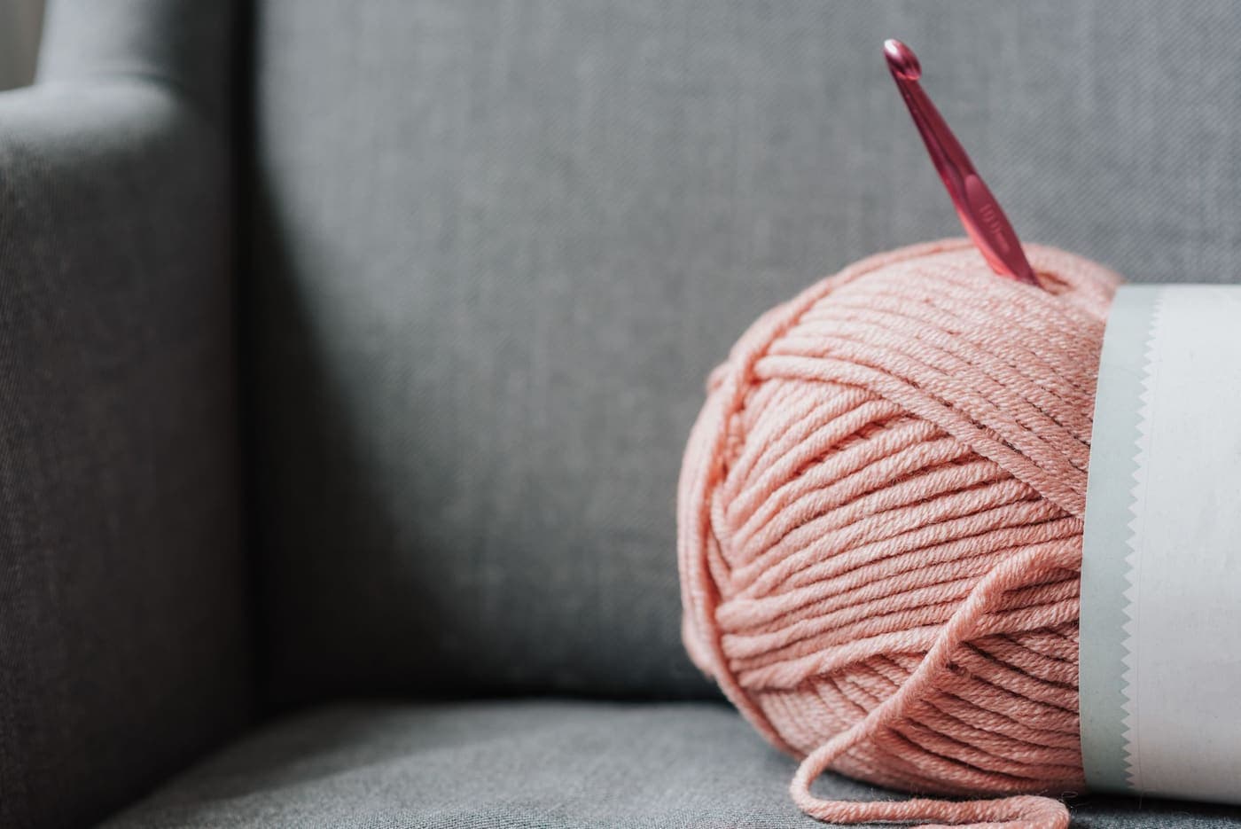 Soft pink yarn and a metal crochet hook resting on a gray couch