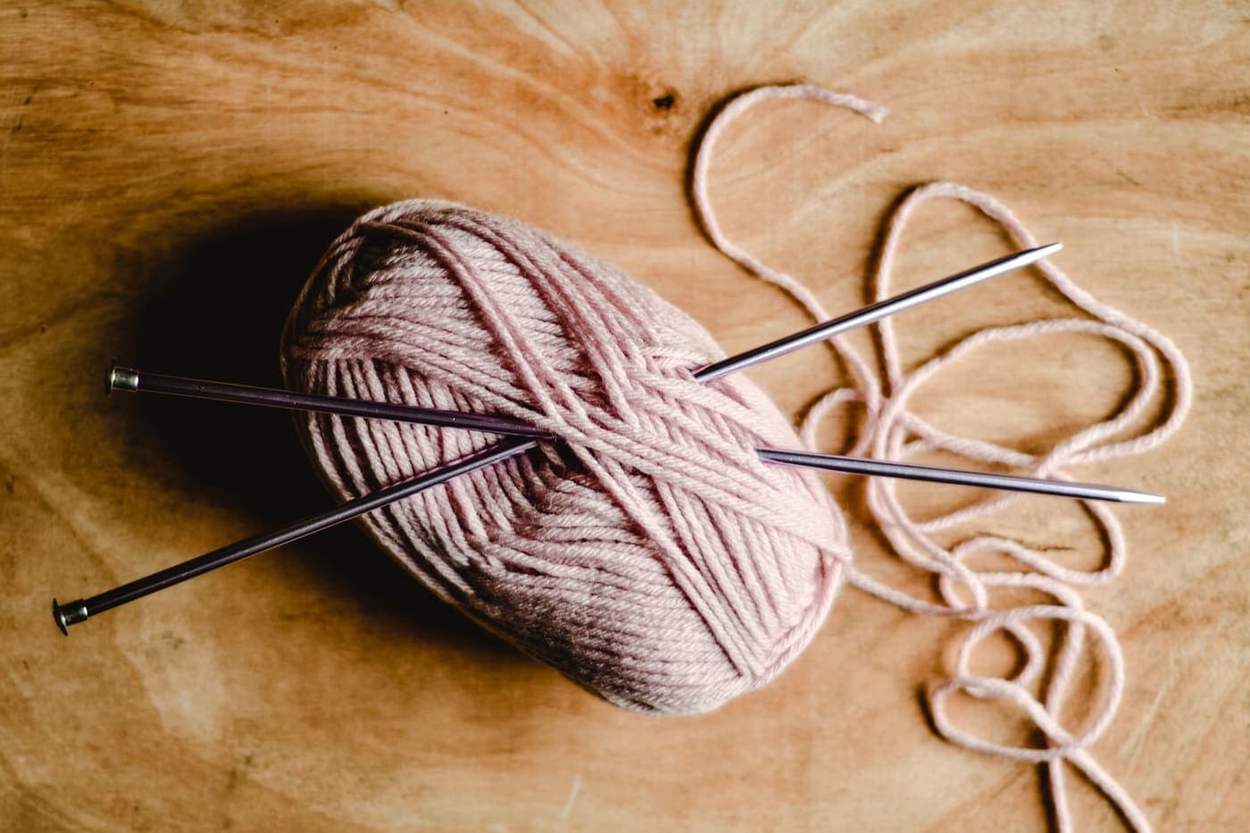 Metal knitting needles pushed into a ball of pink yarn on a wooden surface