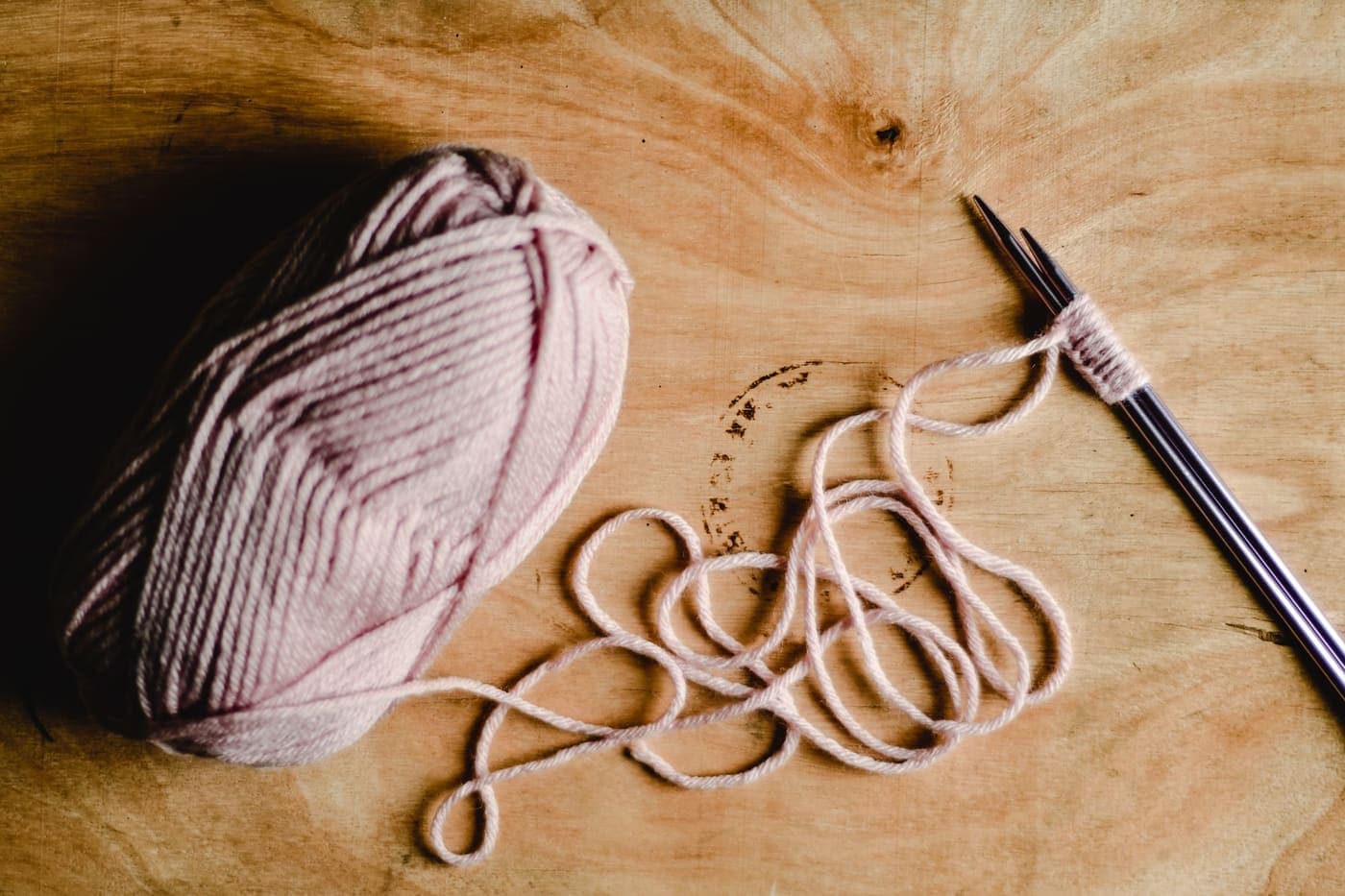A close-up of knitting needles and pink yarn on a wooden surface