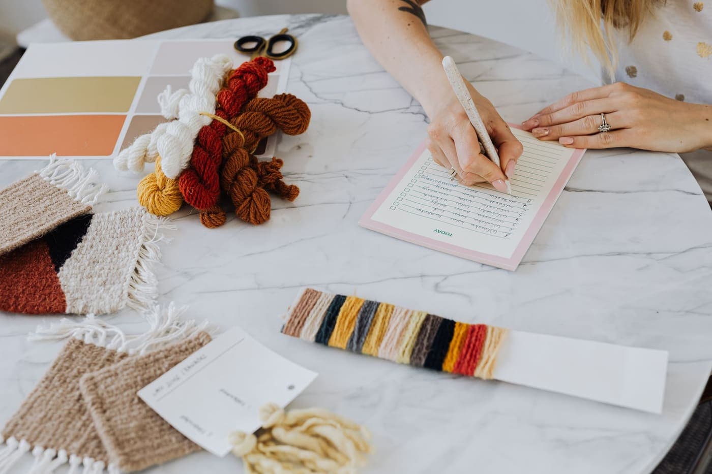 A person writing notes on a marble table beside colorful yarns and swatches