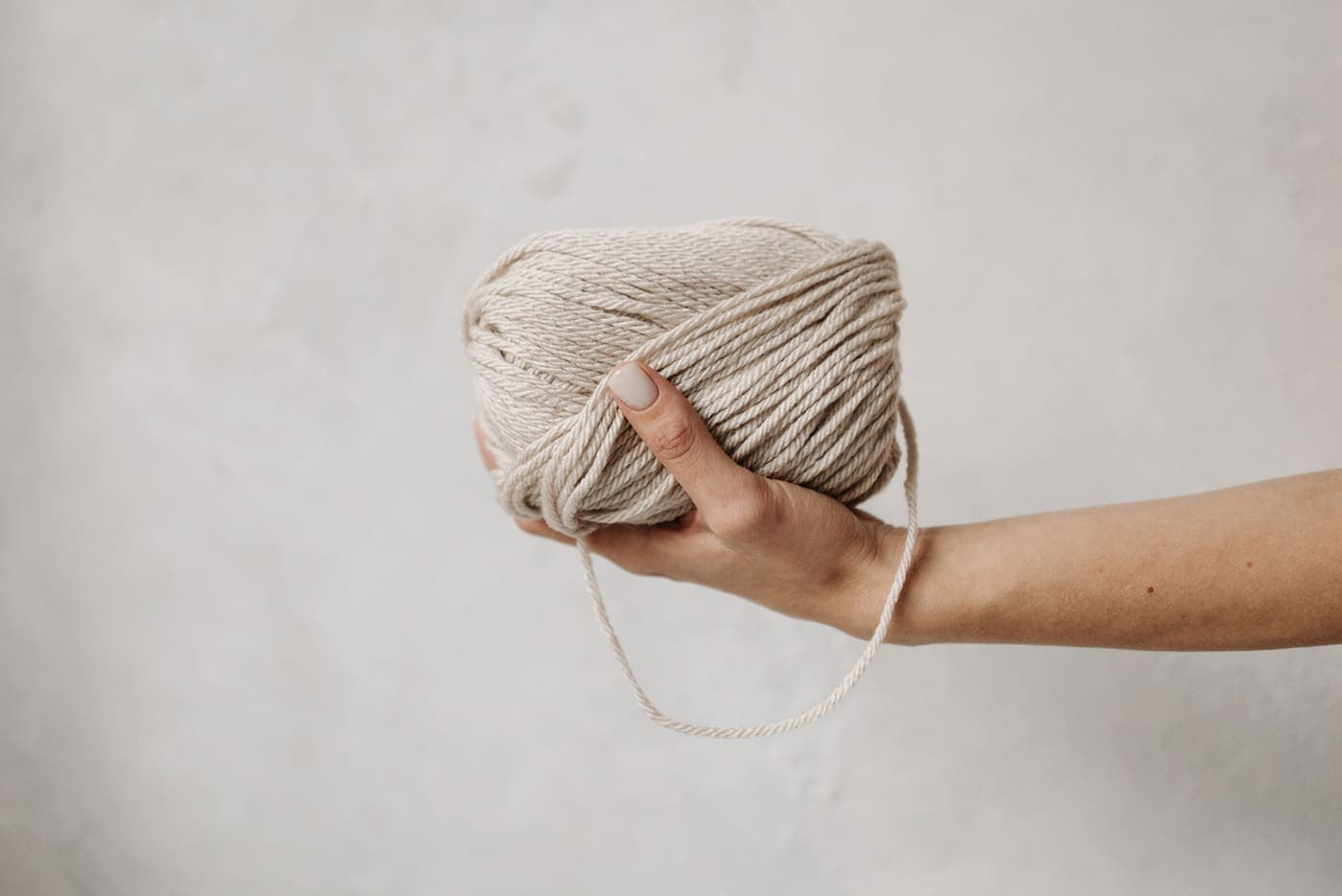 A hand holding a soft beige yarn ball against a neutral background
